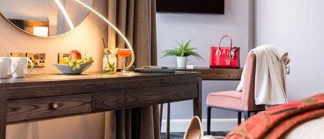 A hotel room with a wooden desk, a mirror, a bowl of fruit, a red handbag, a plant, and a TV mounted on the wall.