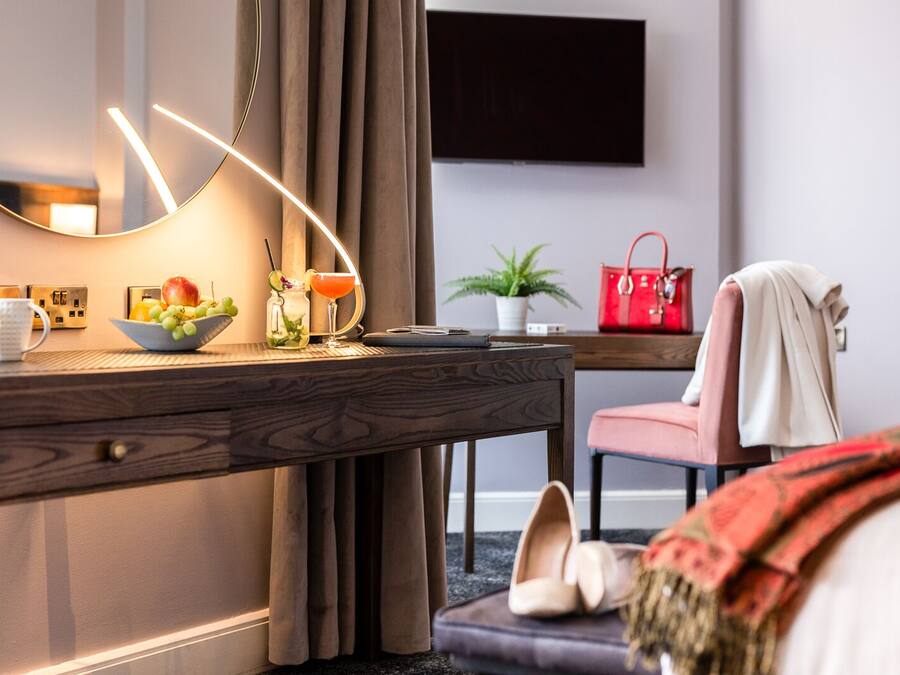 Una habitación de hotel con un escritorio de madera, un espejo, un frutero, un bolso rojo, una planta y un televisor montado en la pared.