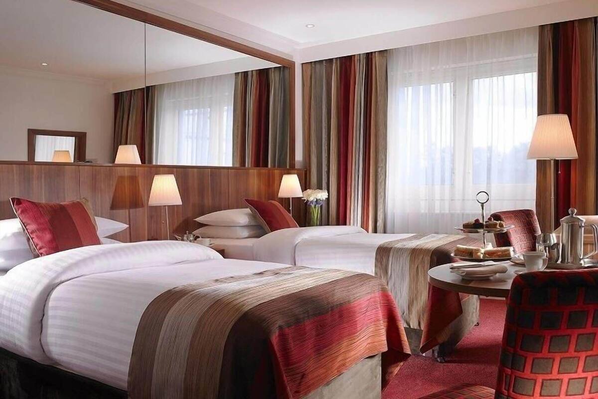 Habitación de hotel con dos camas, una zona de estar con una mesa y un gran ventanal con cortinas.