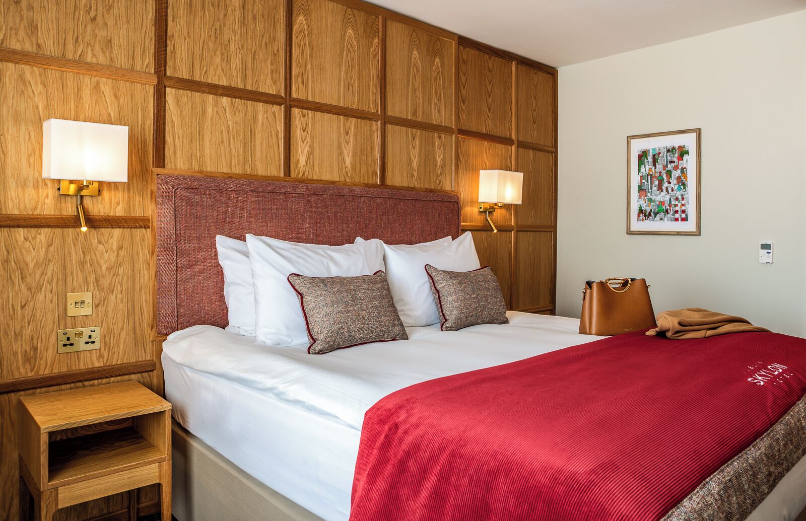 Ein Hotelzimmer mit einer Holzwand, einem großen Bett mit weißen Leinentüchern, einer roten Tagesdecke und zwei Wandlampen.