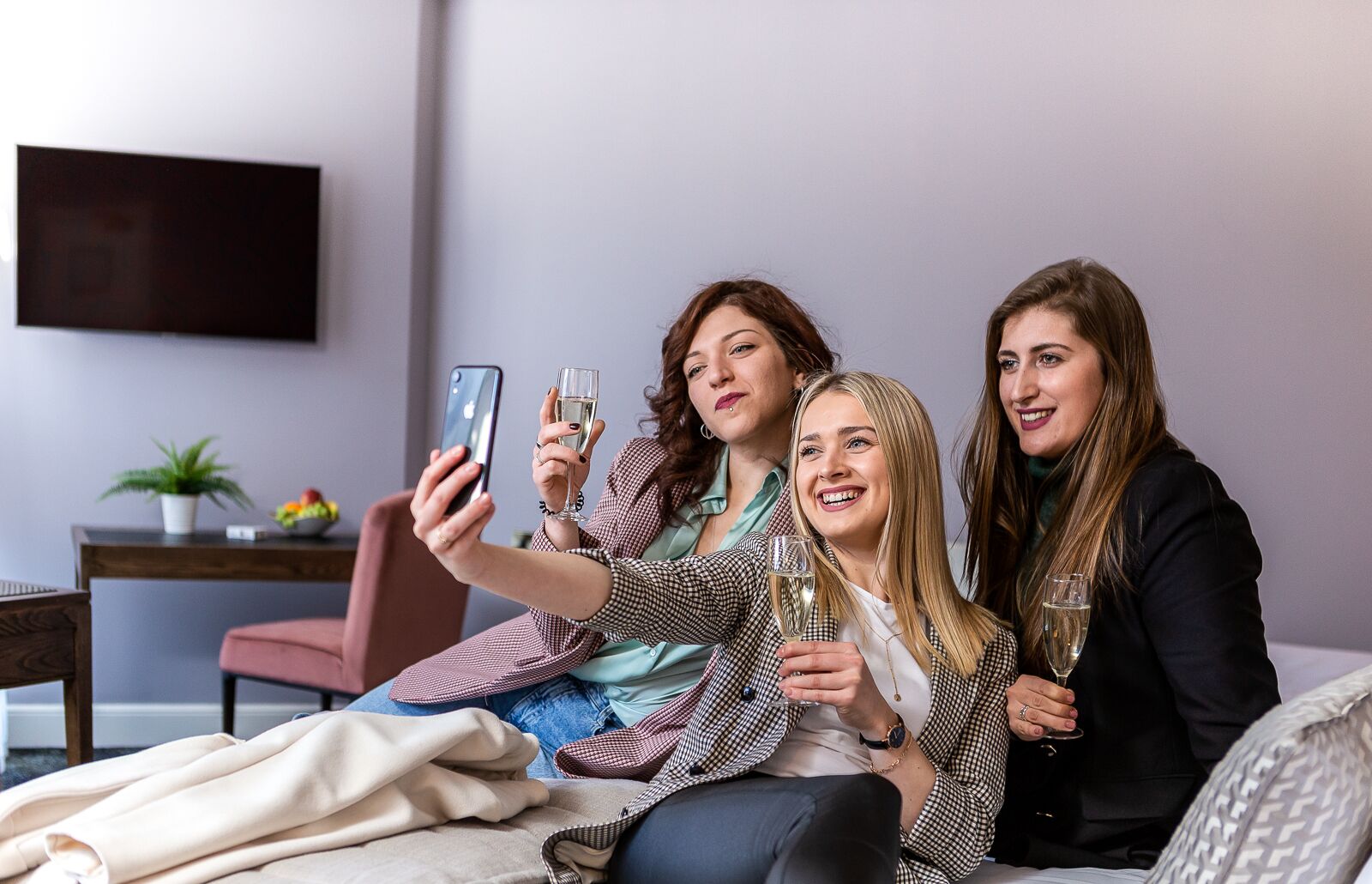 Three people sitting on a bed in a room with a TV on the wall, a plant, and a fruit bowl on a table.
