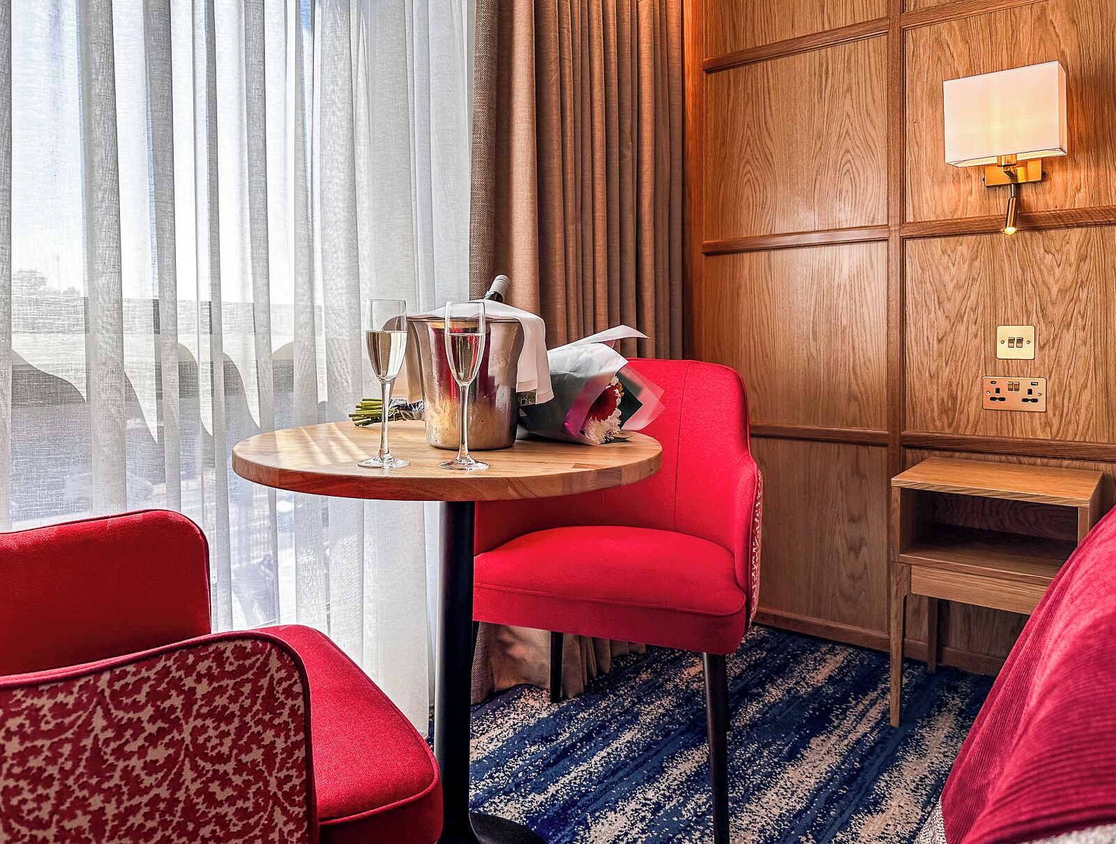 Camera d'albergo con una sedia rossa, un tavolino rotondo con una bottiglia e dei bicchieri, un comodino in legno e vista attraverso tende leggere.