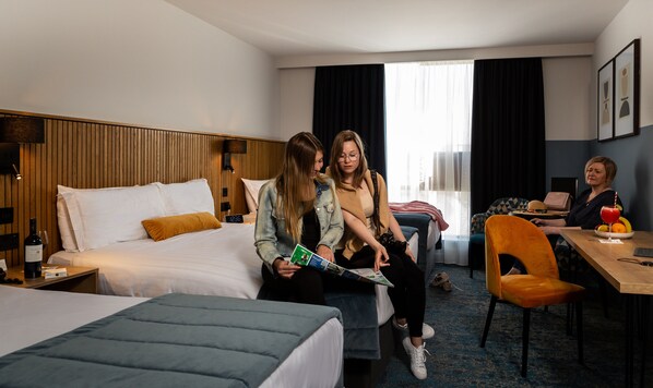 A hotel room with a bed, a desk, and a chair. There is a clock on the wall and a bottle of wine on the nightstand.