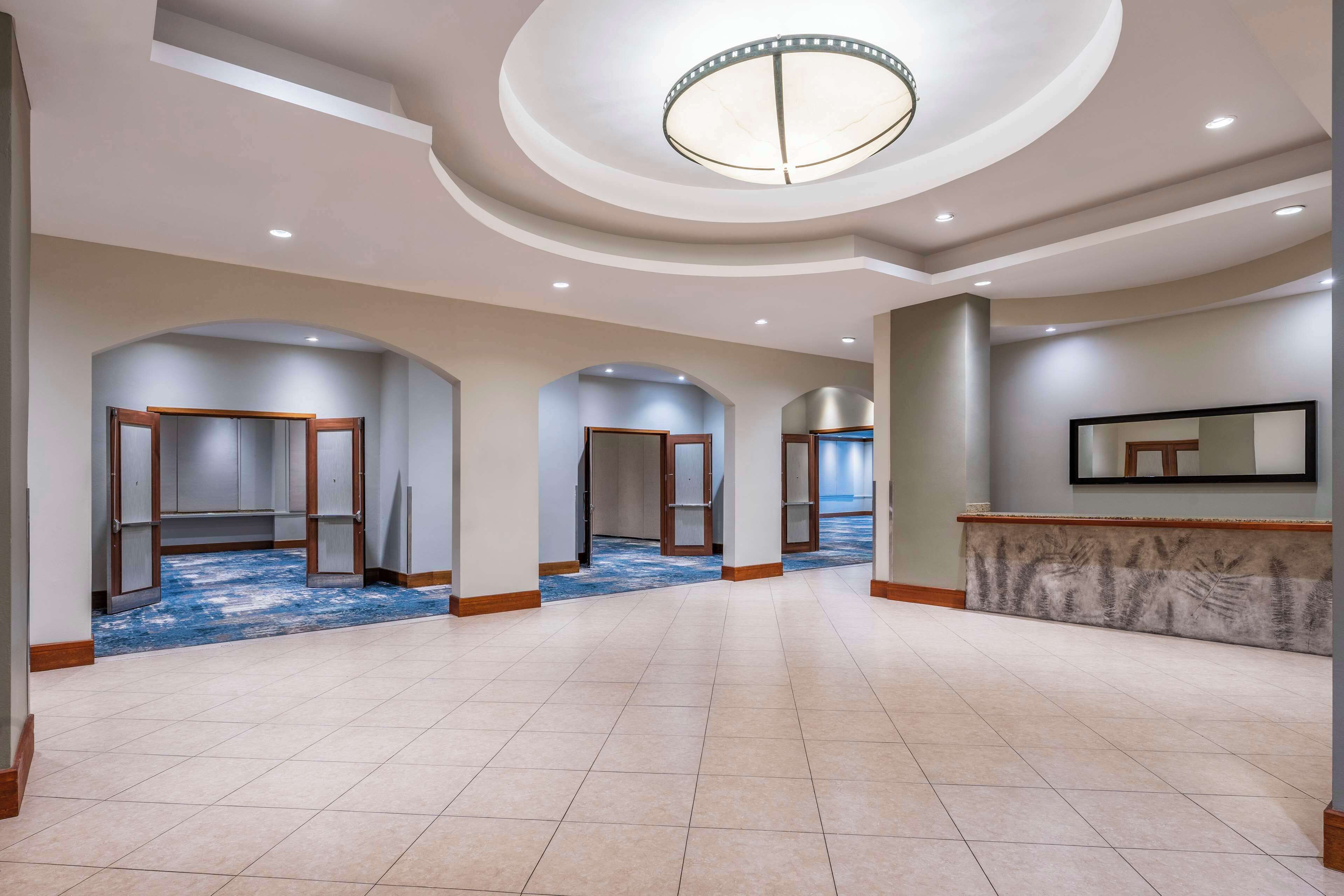 Un hall d’hôtel spacieux avec un plafond haut, des luminaires encastrés et un grand lustre central. Plusieurs portes donnent accès aux chambres, à la réception et au sol recouvert d’un tapis à motifs.