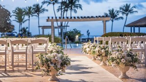 Un pasillo nupcial a la orilla de la playa con sillas blancas y arreglos florales, palmeras y un cielo despejado.
