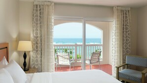 Habitación de hotel con cama, mesita de noche, lámpara, silla y balcón con vista al océano.