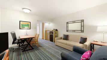 Una habitación de hotel con escritorio, dos sofás, televisor, mesa de comedor y zona de cocina.