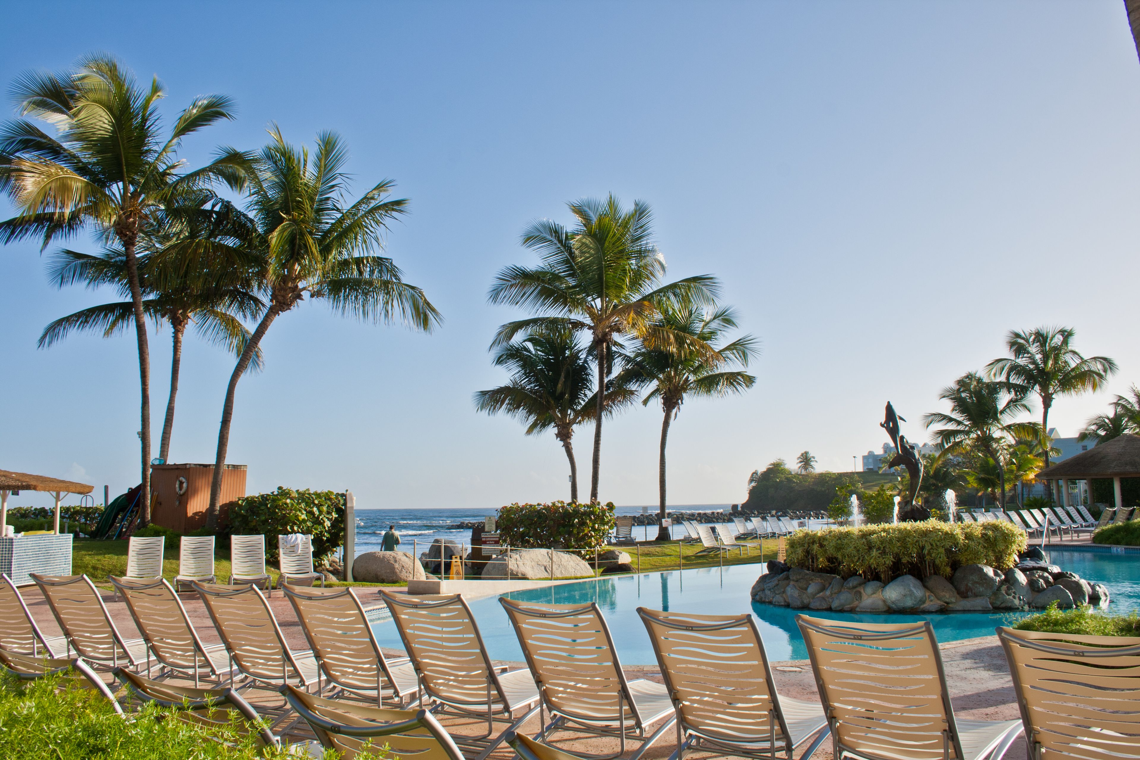 A poolside area with lounge chairs, palm trees, and a view of the ocean.