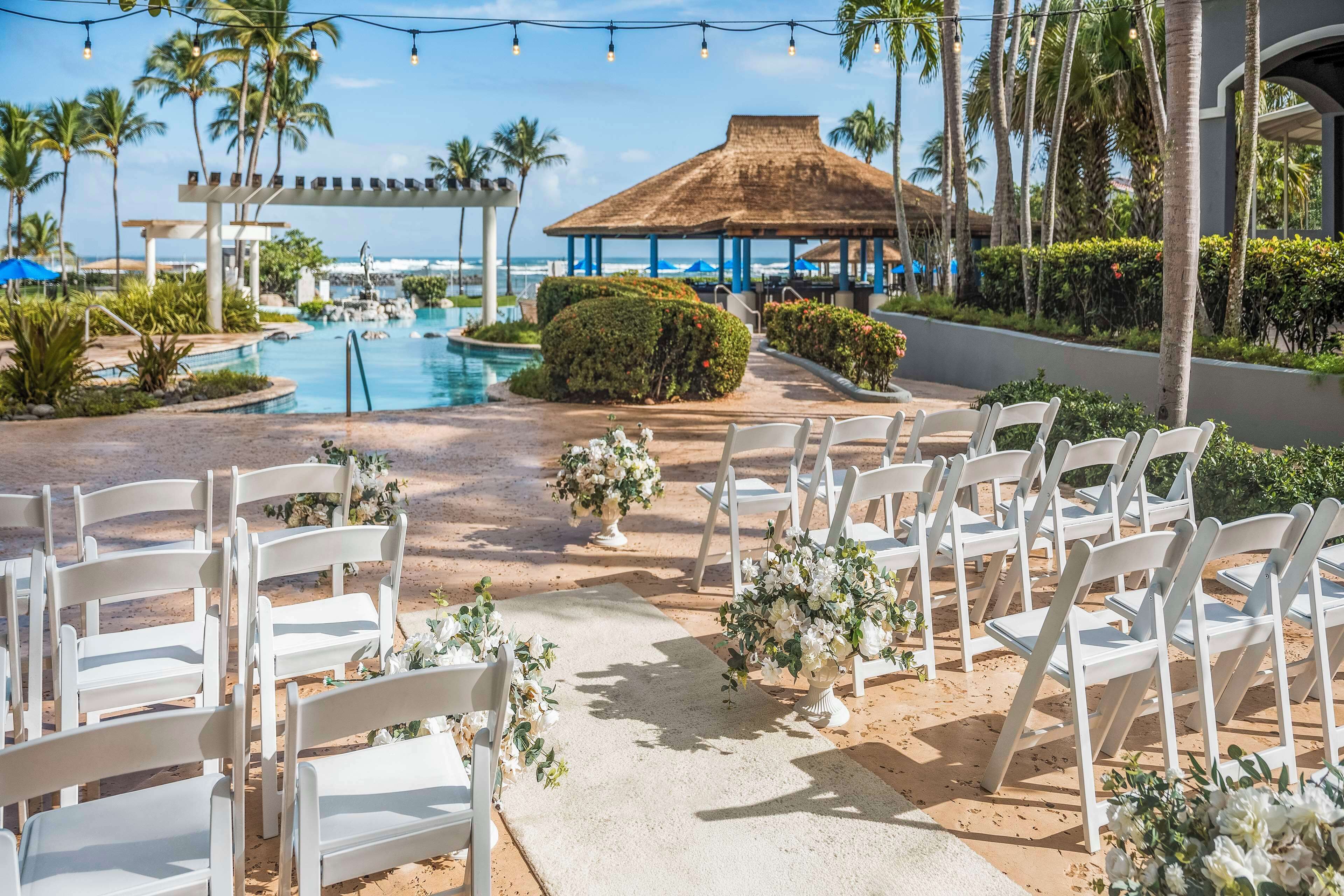 Preparação para uma cerimônia de casamento, com cadeiras brancas e decoração floral em uma área arenosa, com piscina e palmeiras ao fundo.