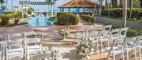 Preparação para uma cerimônia de casamento, com cadeiras brancas e decoração floral em uma área arenosa, com piscina e palmeiras ao fundo.