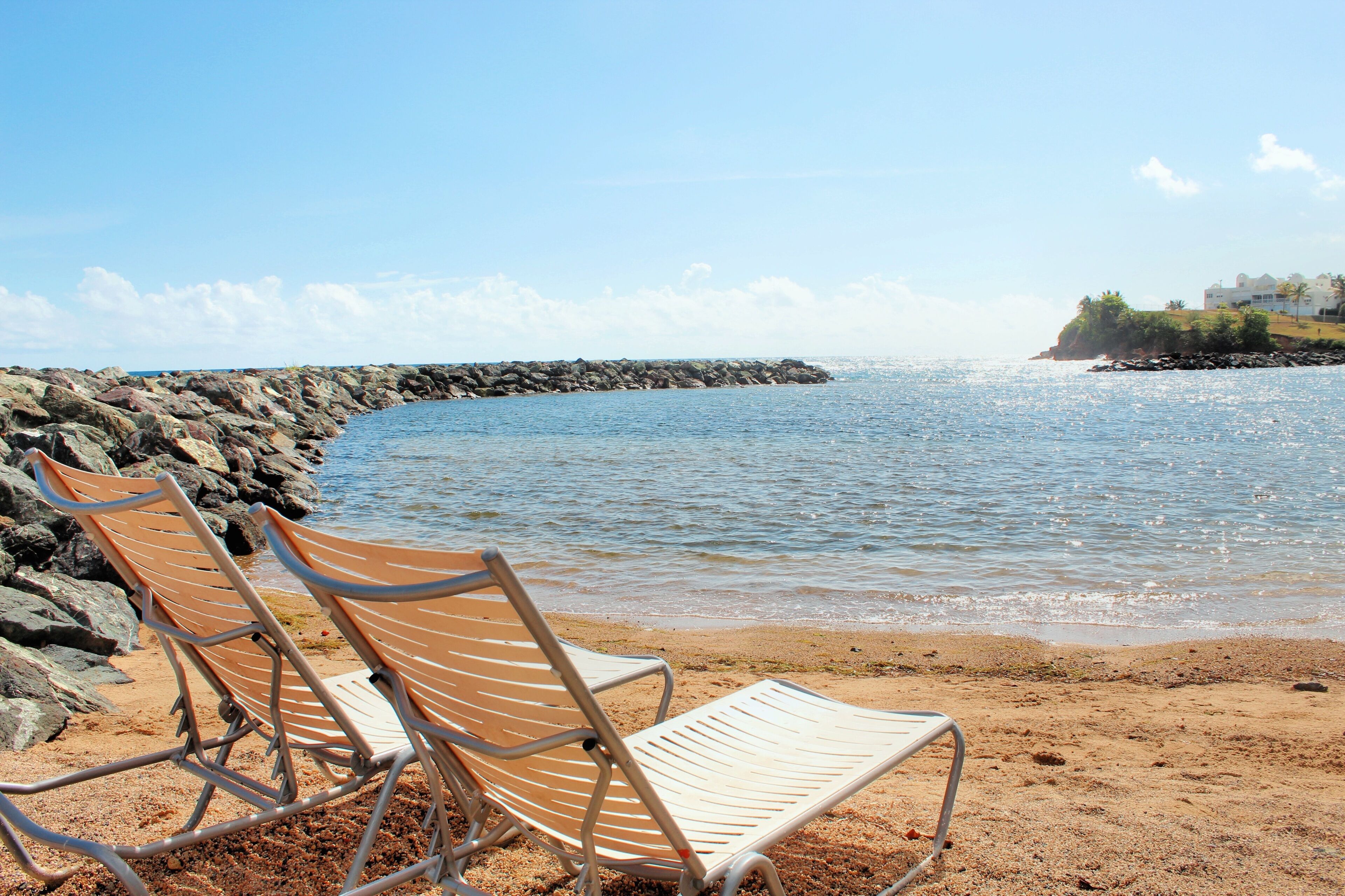 Two beach chairs on a sandy shore with a rocky breakwater and calm sea in the background.