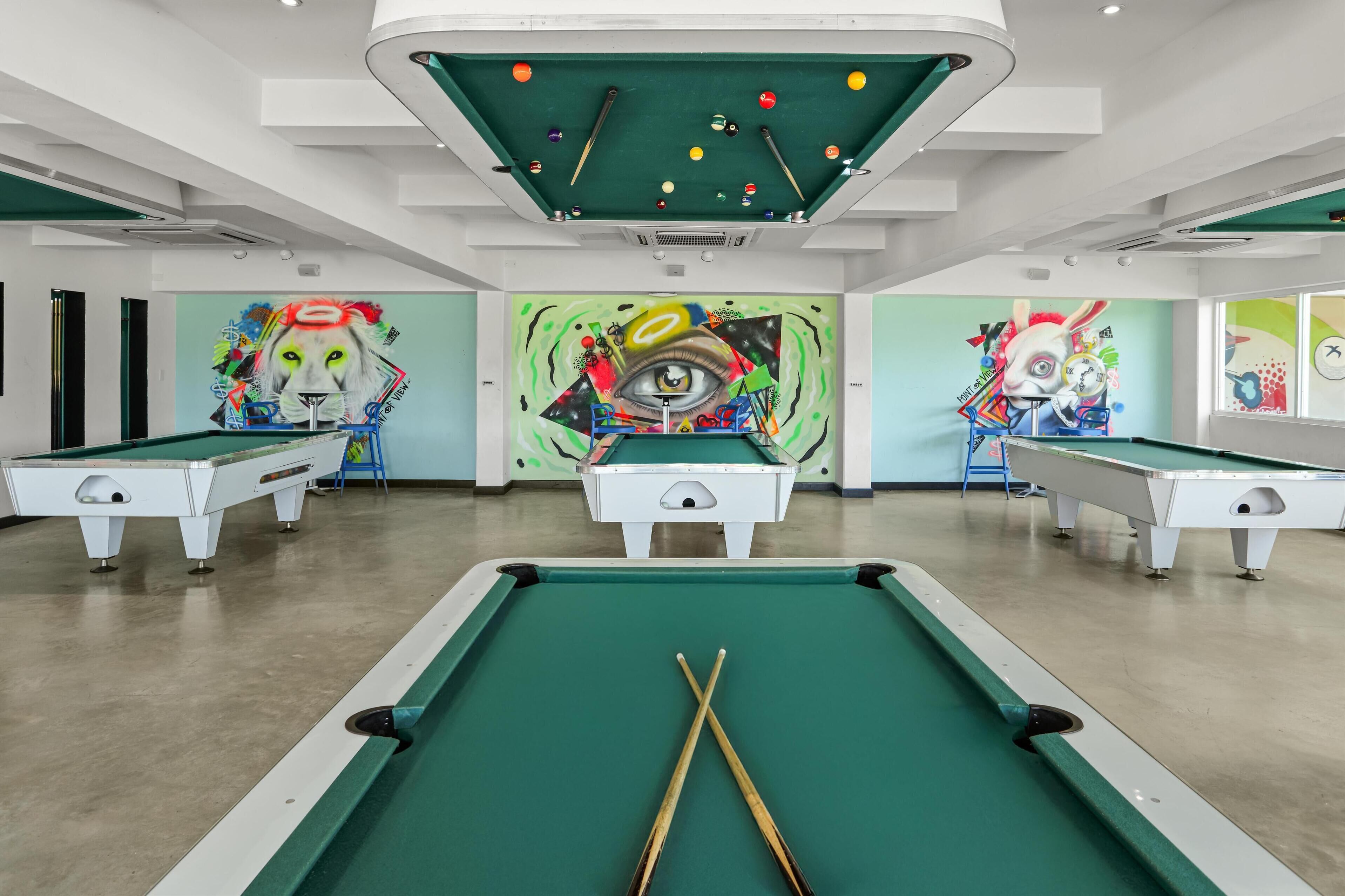 A pool hall with multiple pool tables, a green felt table in the foreground, and colorful murals on the walls.