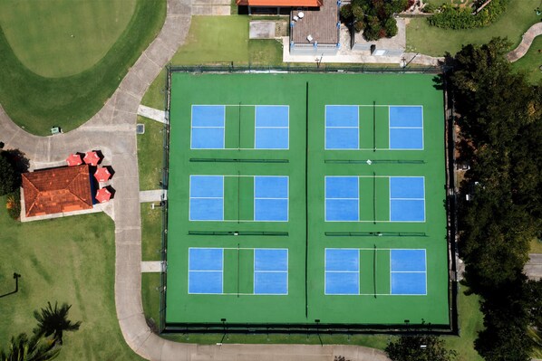 Vista aérea de un complejo deportivo con varias canchas de tenis, una zona de asientos techada y zonas verdes circundantes.
