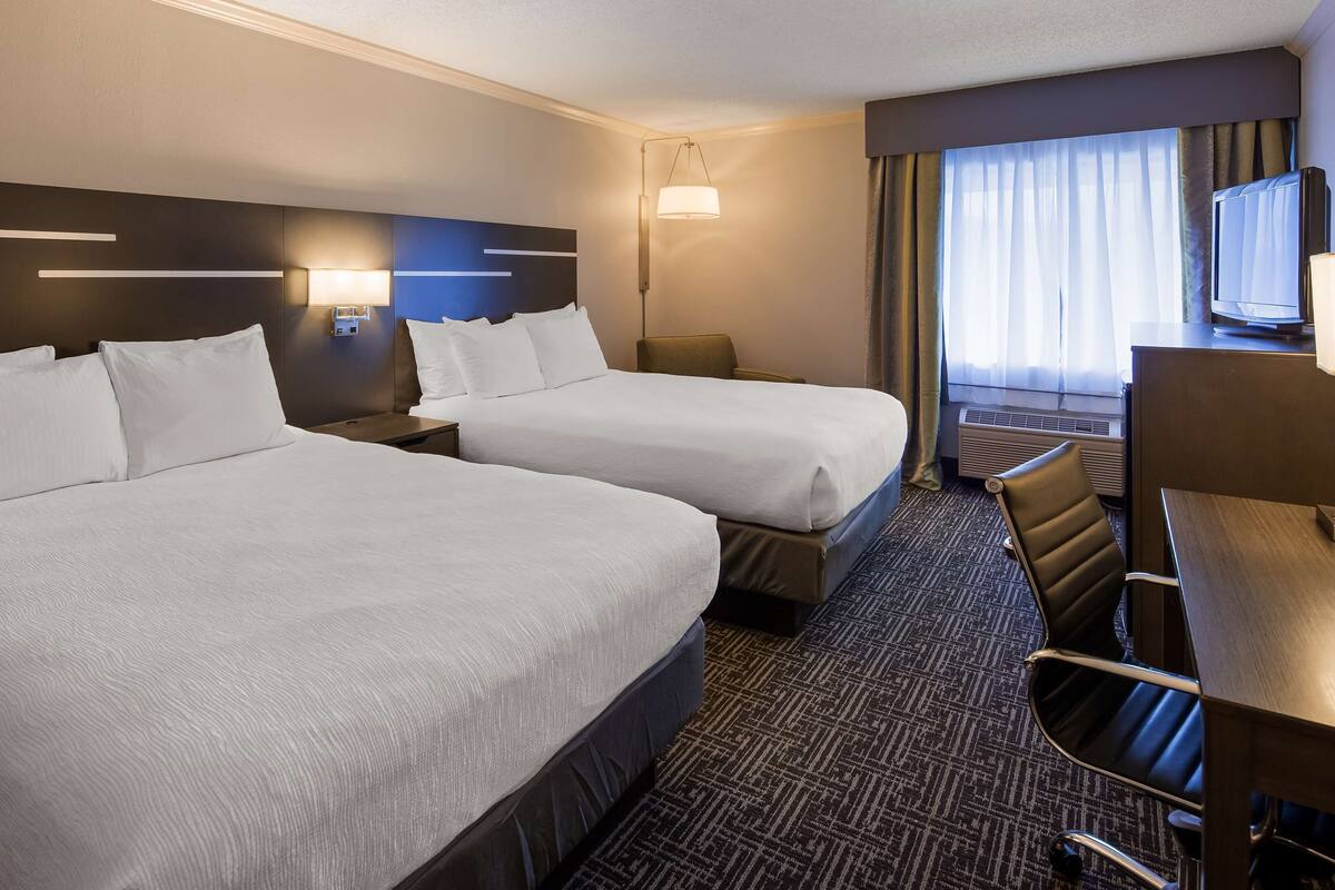 A hotel room with two beds, a desk, a chair, a TV, and a window with curtains.