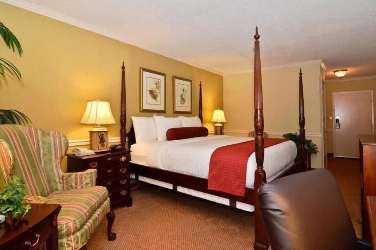 A four-poster bed with a red throw, a striped armchair, a wooden nightstand, and two wall-mounted lamps.