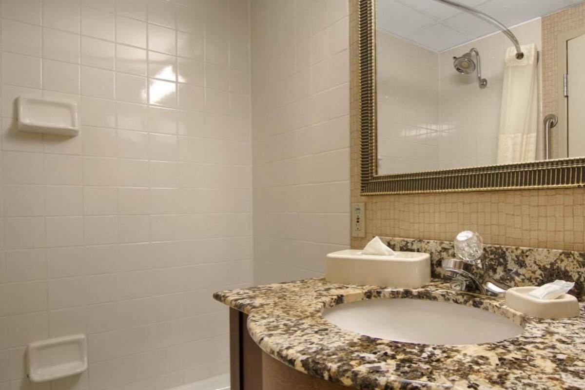 A bathroom with a white tiled wall, a granite countertop sink, a mirror with a gold frame, and a shower area.