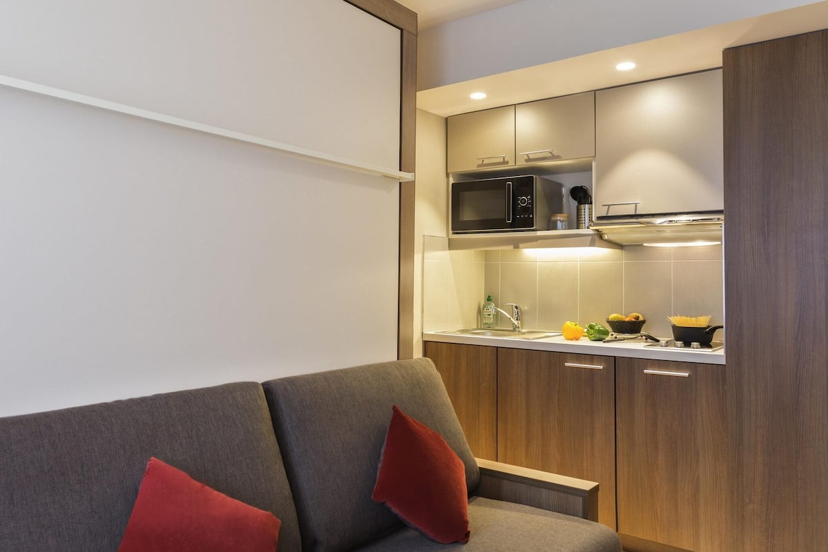 Eine moderne Küche mit Mikrowelle, Herd und Spüle. Im Vordergrund steht ein graues Sofa mit roten Kissen.