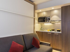 Eine moderne Küche mit Mikrowelle, Herd und Spüle. Im Vordergrund steht ein graues Sofa mit roten Kissen.