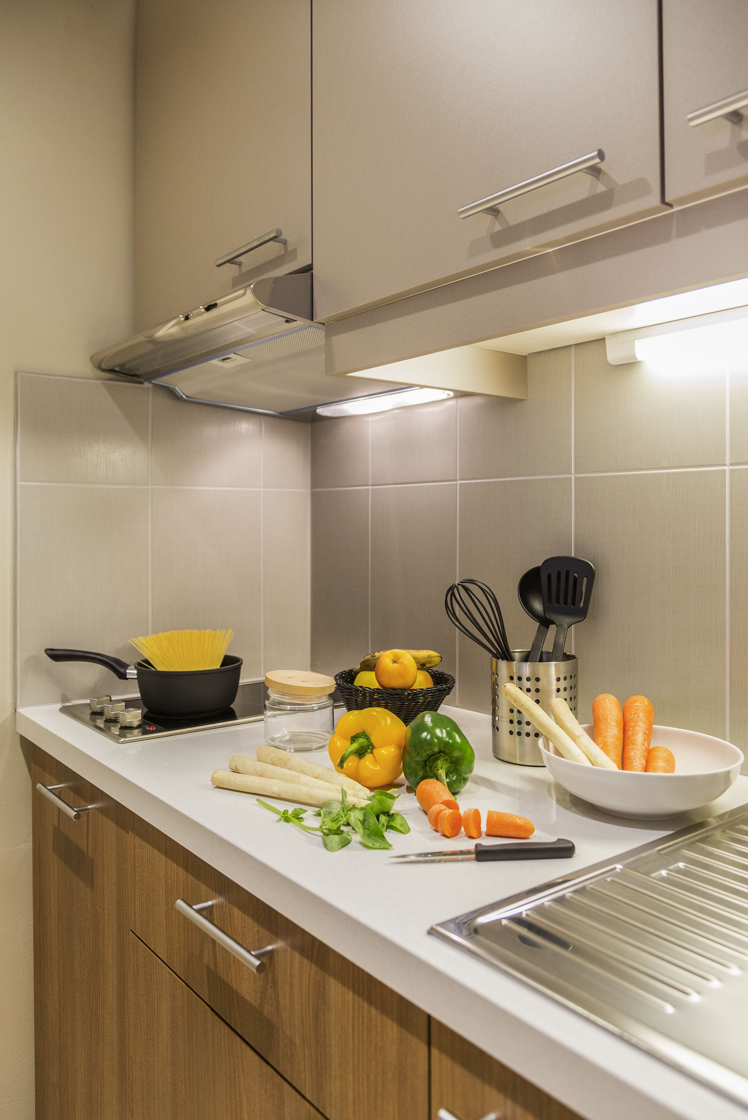 A kitchen with a stove, pots, vegetables, and utensils on the counter.
