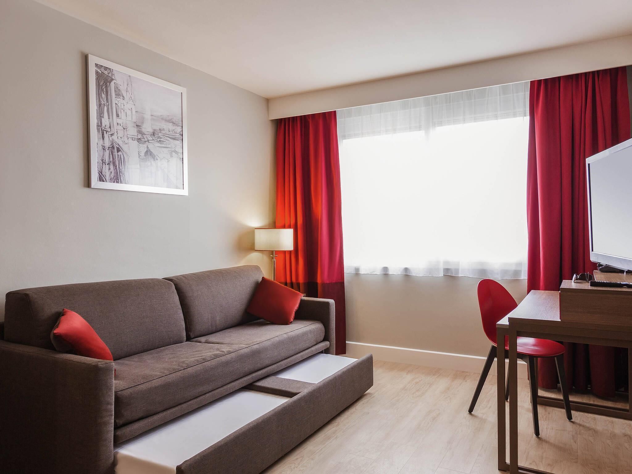 Een moderne hotelkamer met een bank, een bureau, een stoel, een lamp en een groot raam met rode gordijnen.