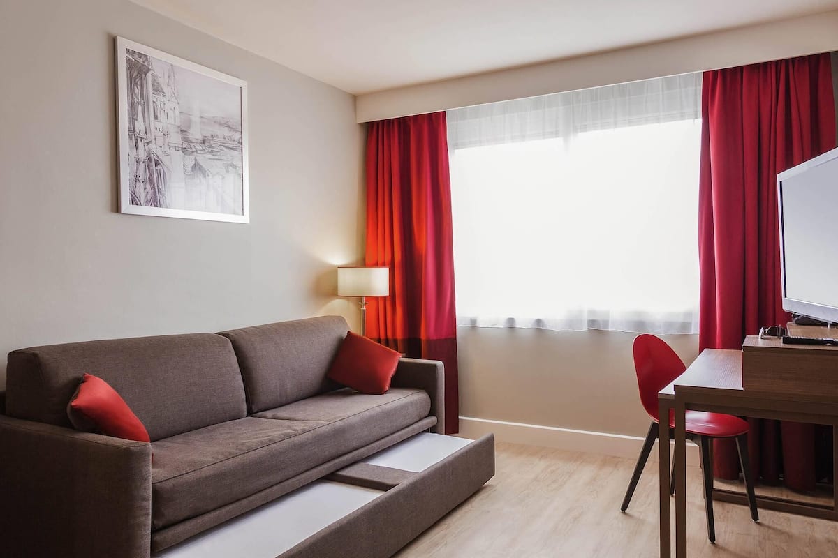 A modern hotel room with a sofa, a desk, a chair, a lamp, and a large window with red curtains.