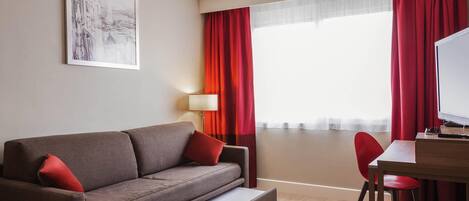 Habitación de hotel moderna con sofá, escritorio, silla, lámpara y un amplio ventanal con cortinas rojas.