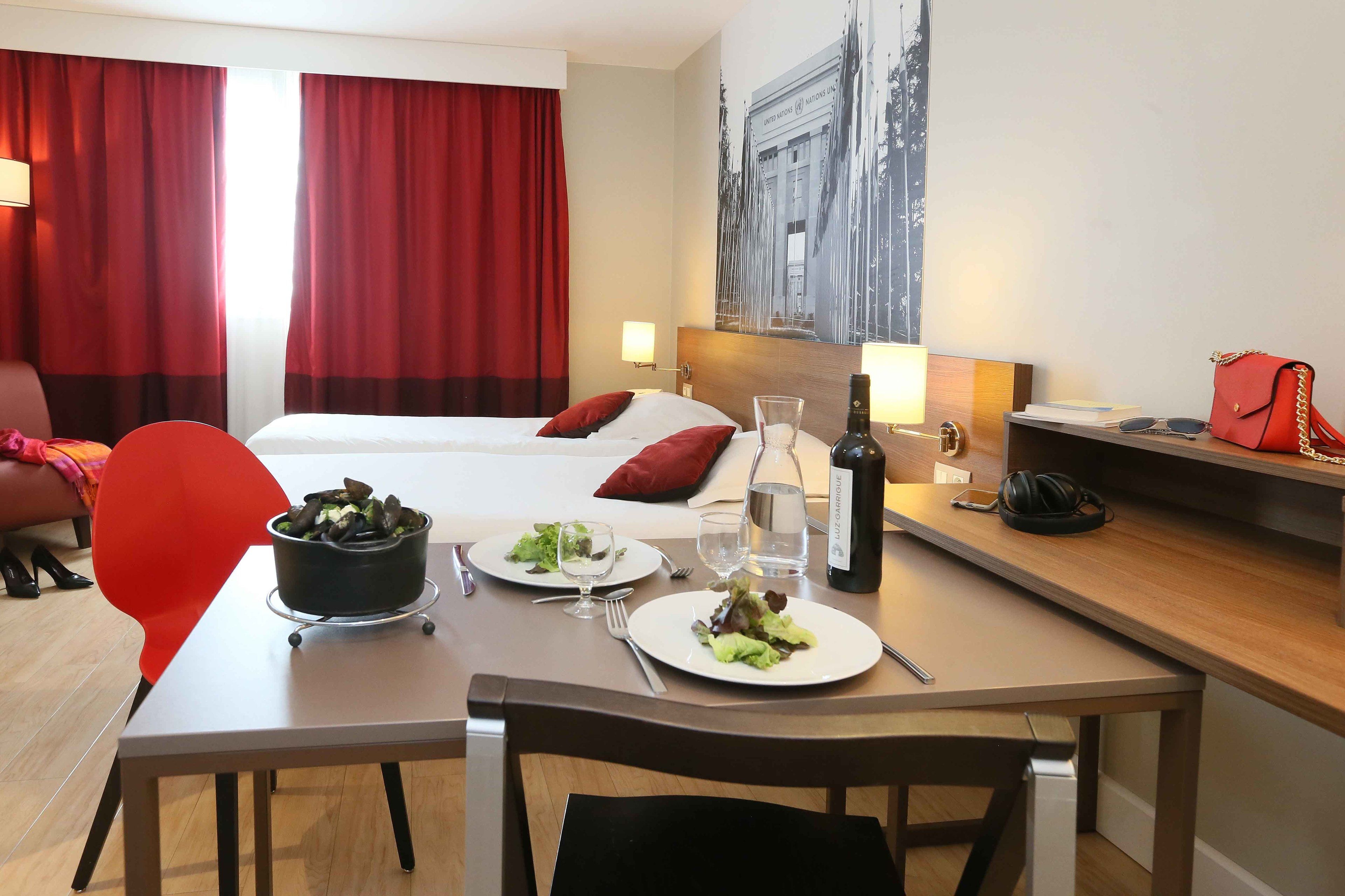 A hotel room with a bed, a dining table with plates of food, a bottle of wine, and a red handbag on a side table.