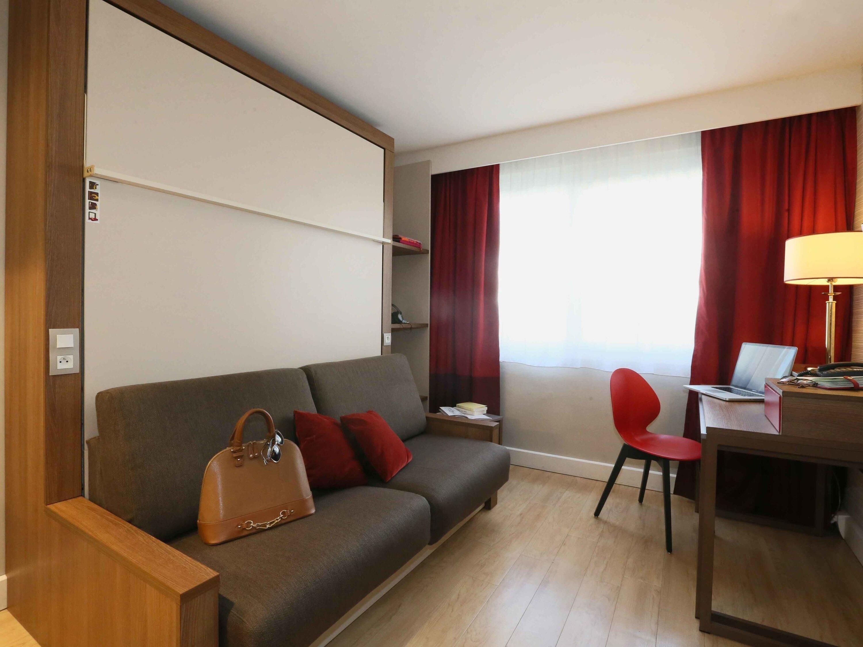 A hotel room with a sofa, a desk with a laptop, a chair, and a lamp.