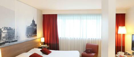 A hotel room with a large bed, a chair, a desk with a lamp, and a television.
