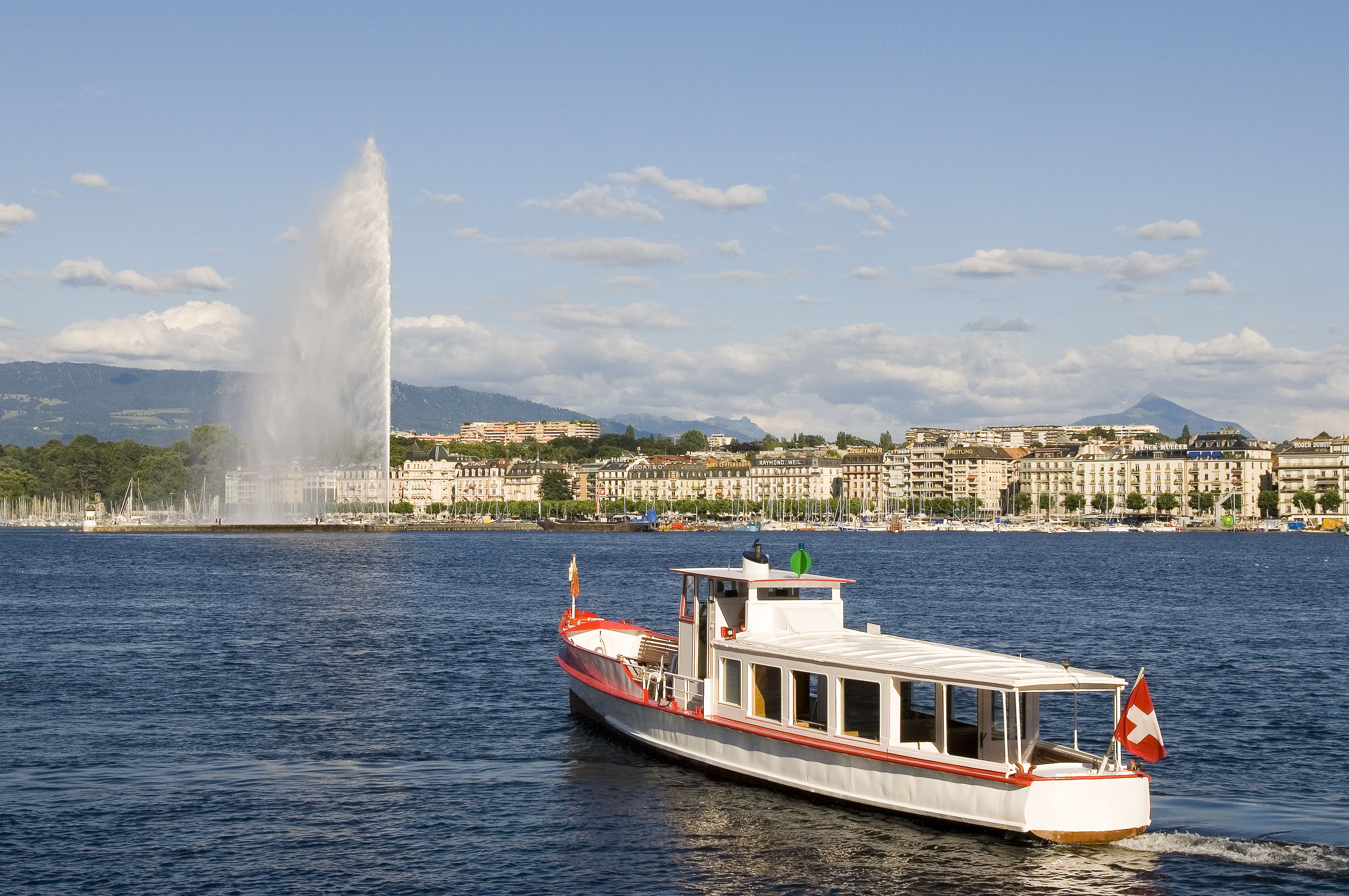 A boat with a flag on a calm water body with a fountain and buildings in the background.