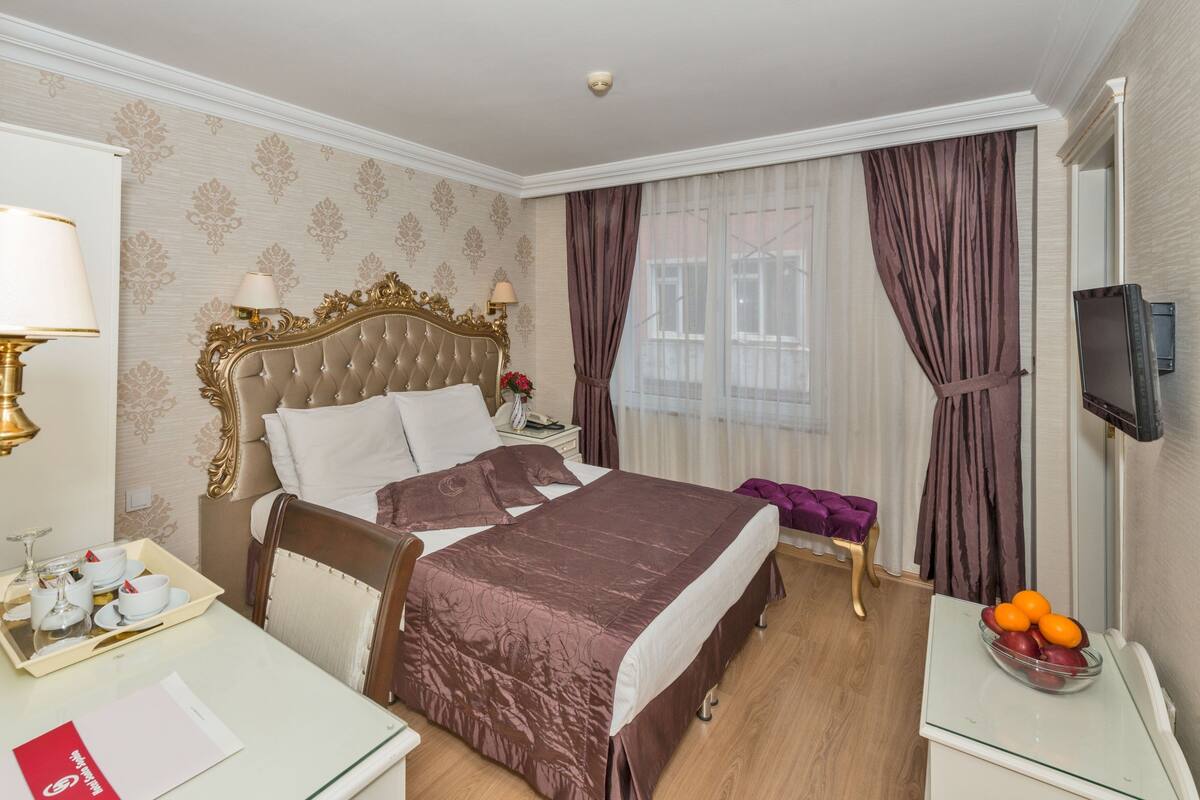 A hotel room with a large bed, a desk with a tray of breakfast items, a bedside table with a vase of flowers, and a television on the wall.