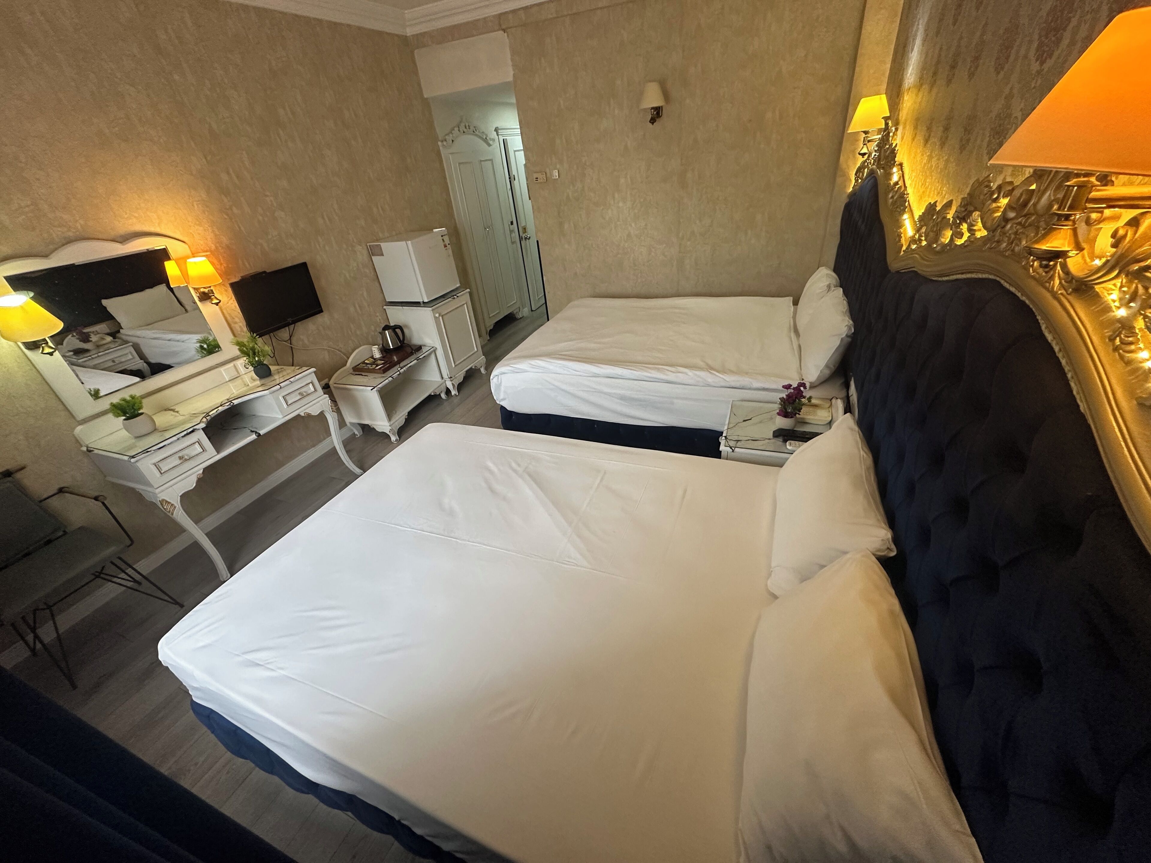 Habitación de hotel con dos camas, un escritorio con lámpara, un espejo, un armario y televisión.