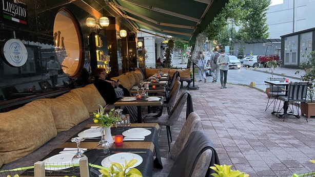 Ein Restaurant im Freien mit Tischen und Bänken, einer grünen Markise und einem Schild mit der Aufschrift 'Lugano'.
