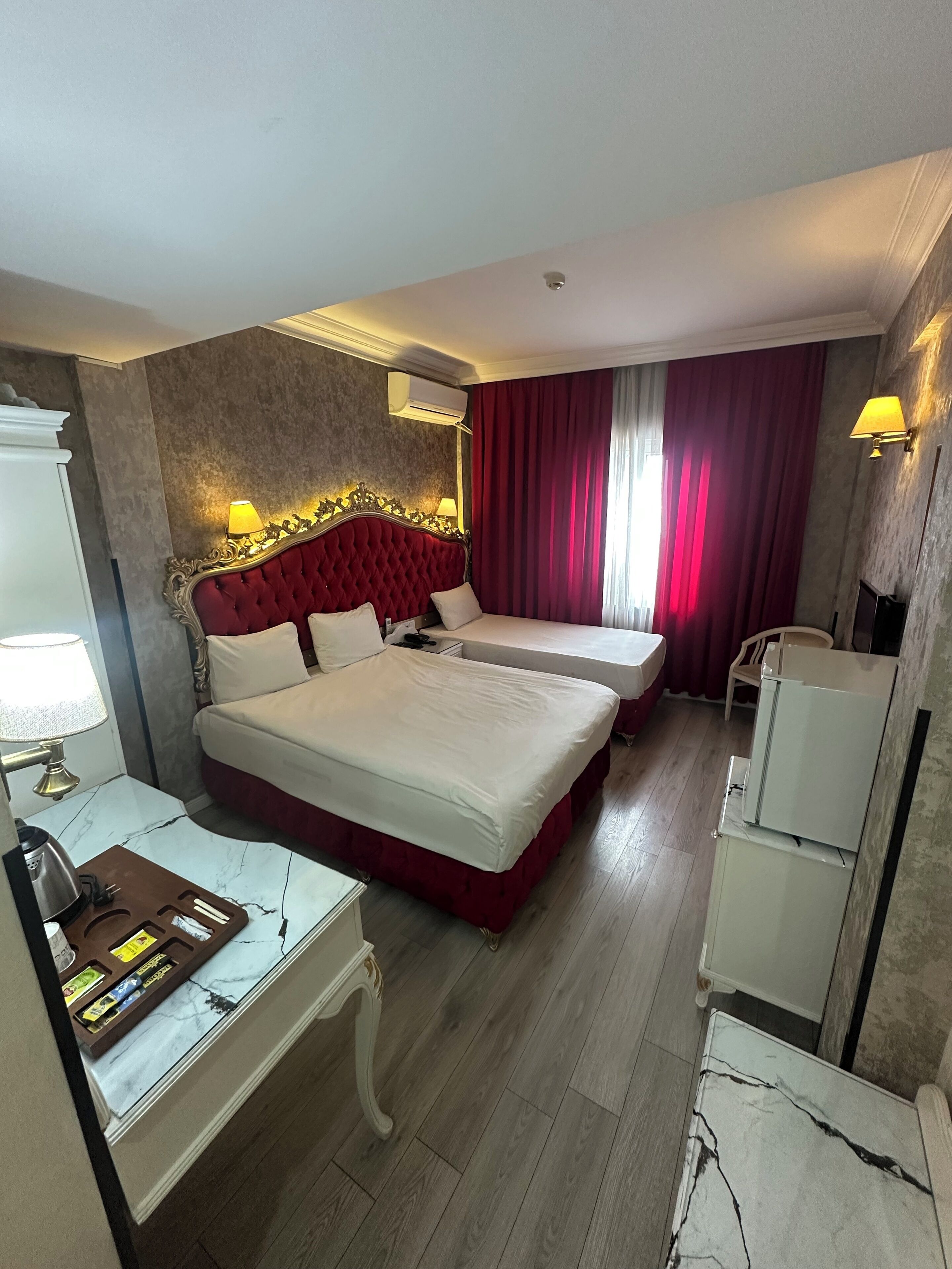 A hotel room with two beds, a desk with a kettle, a chair, and a small refrigerator.