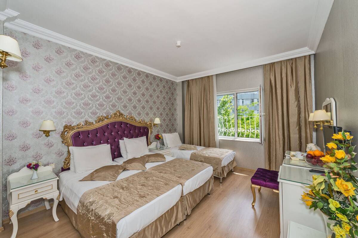 A hotel room with two beds, a vanity with a mirror, a window with a view of greenery, and a nightstand with a lamp and flowers.