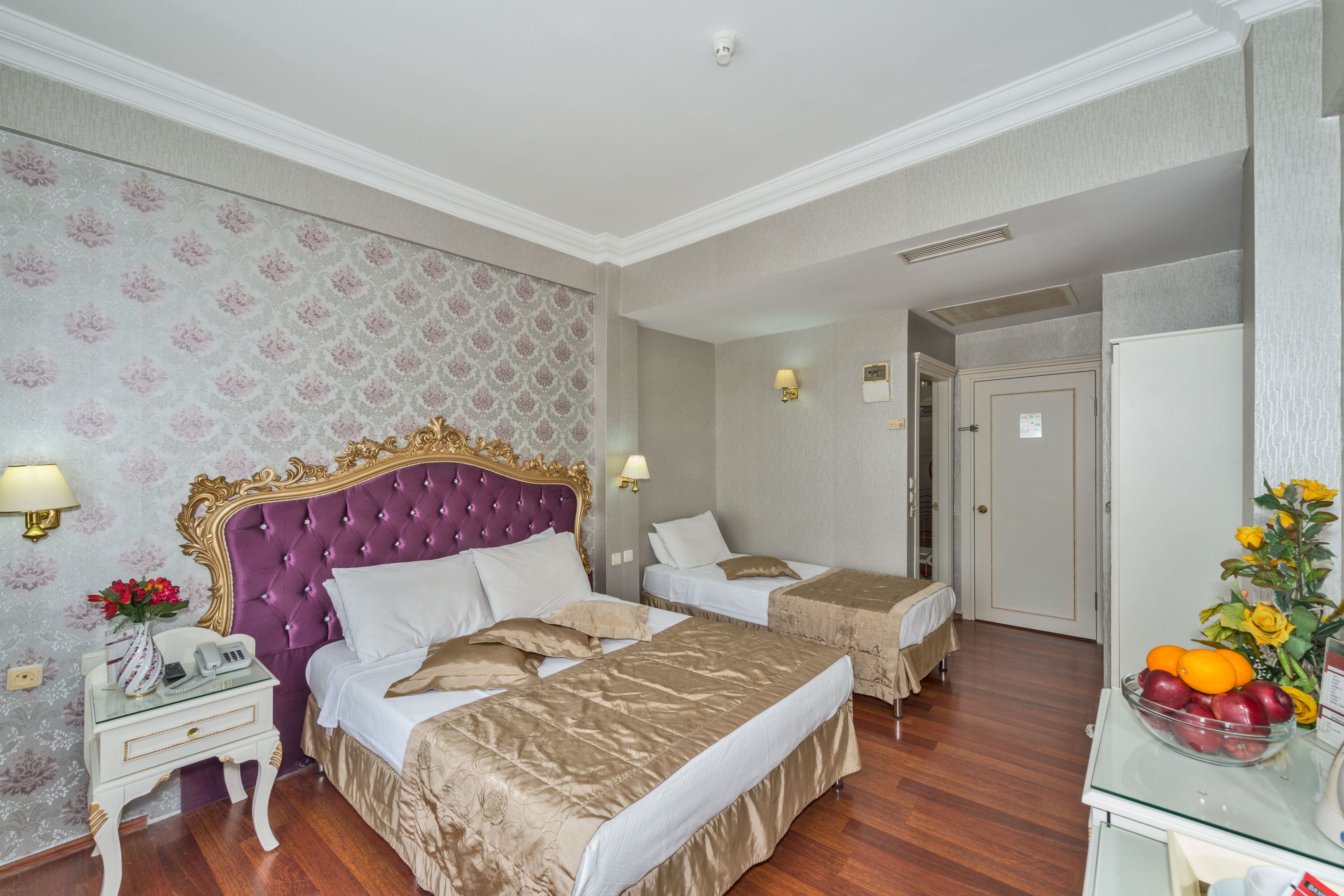 Une chambre d’hôtel avec deux lits, une coiffeuse avec un miroir, une petite table ornée d’un vase de fleurs et un panier de fruits posé à côté.