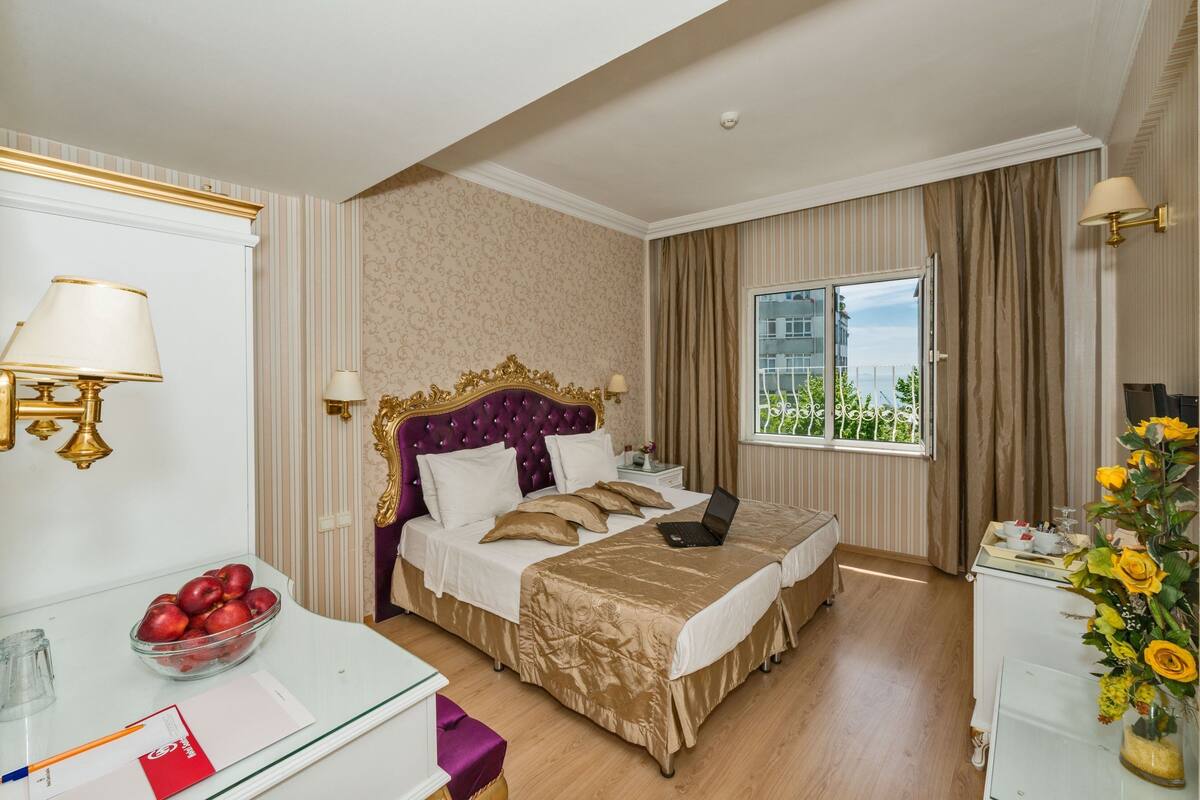 A hotel room with a large bed, a desk with a laptop, a bowl of fruit, and a window with a view of buildings.
