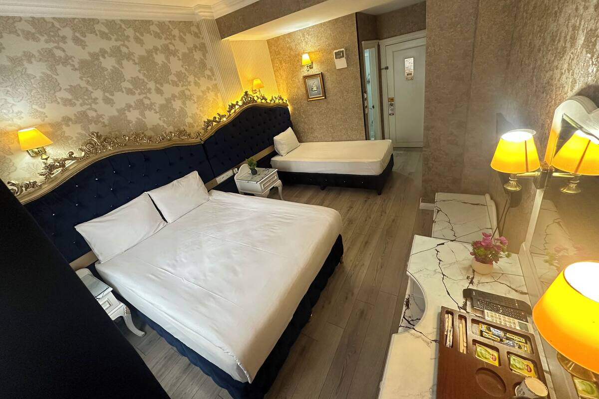 A hotel room with a large bed, a smaller bed, a desk with a lamp, and a minibar.