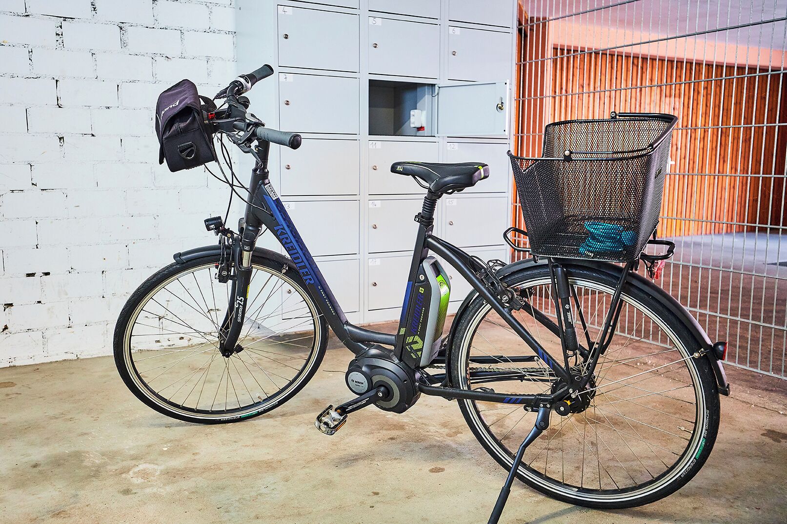 A stationary bicycle with a basket and a bag attached to the handlebars, parked against a white brick wall with a metal fence in the background.