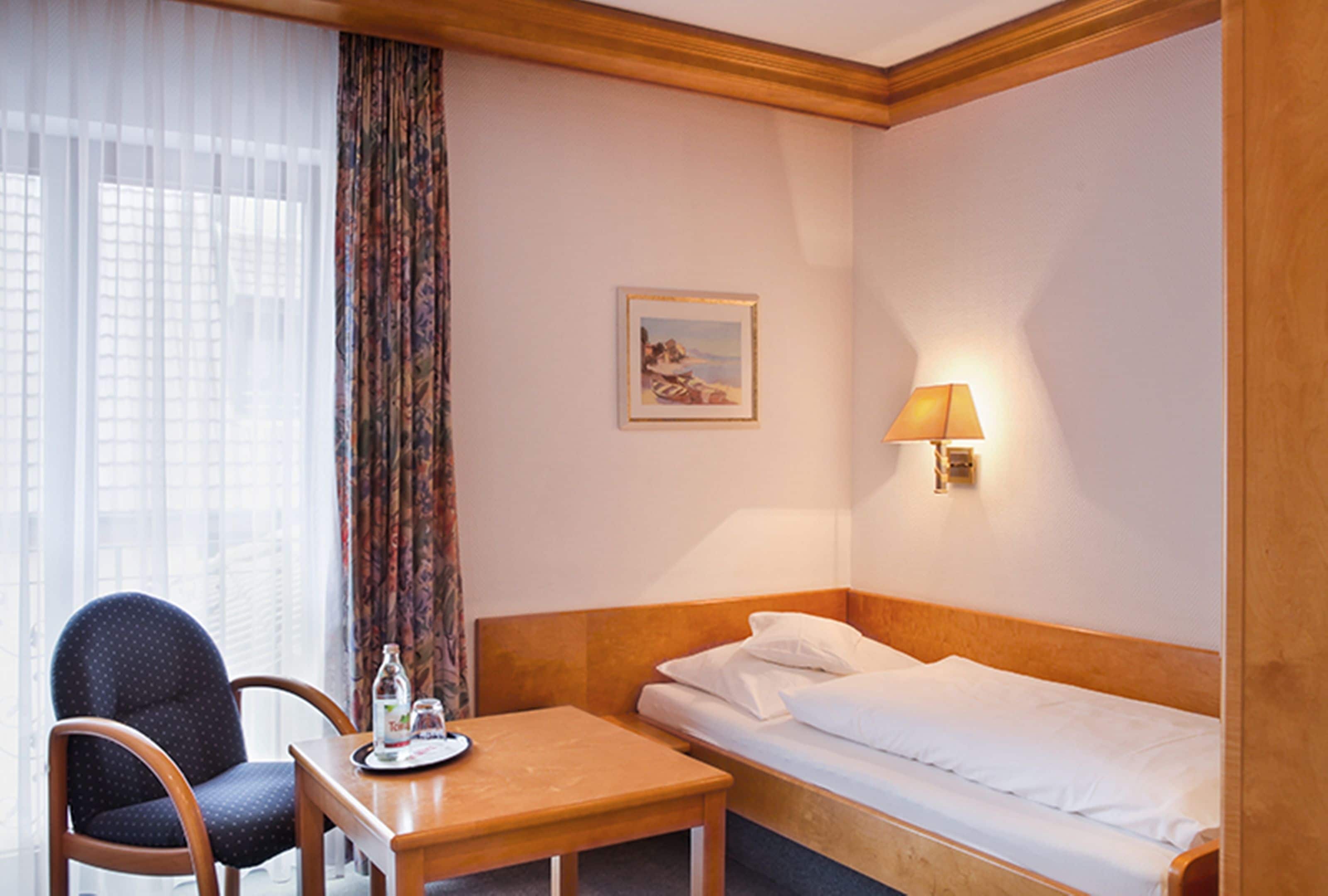 A hotel room with a single bed, a wooden headboard, a small table with a bottle and glass, a chair, a window with sheer curtains, and a wall-mounted lamp.
