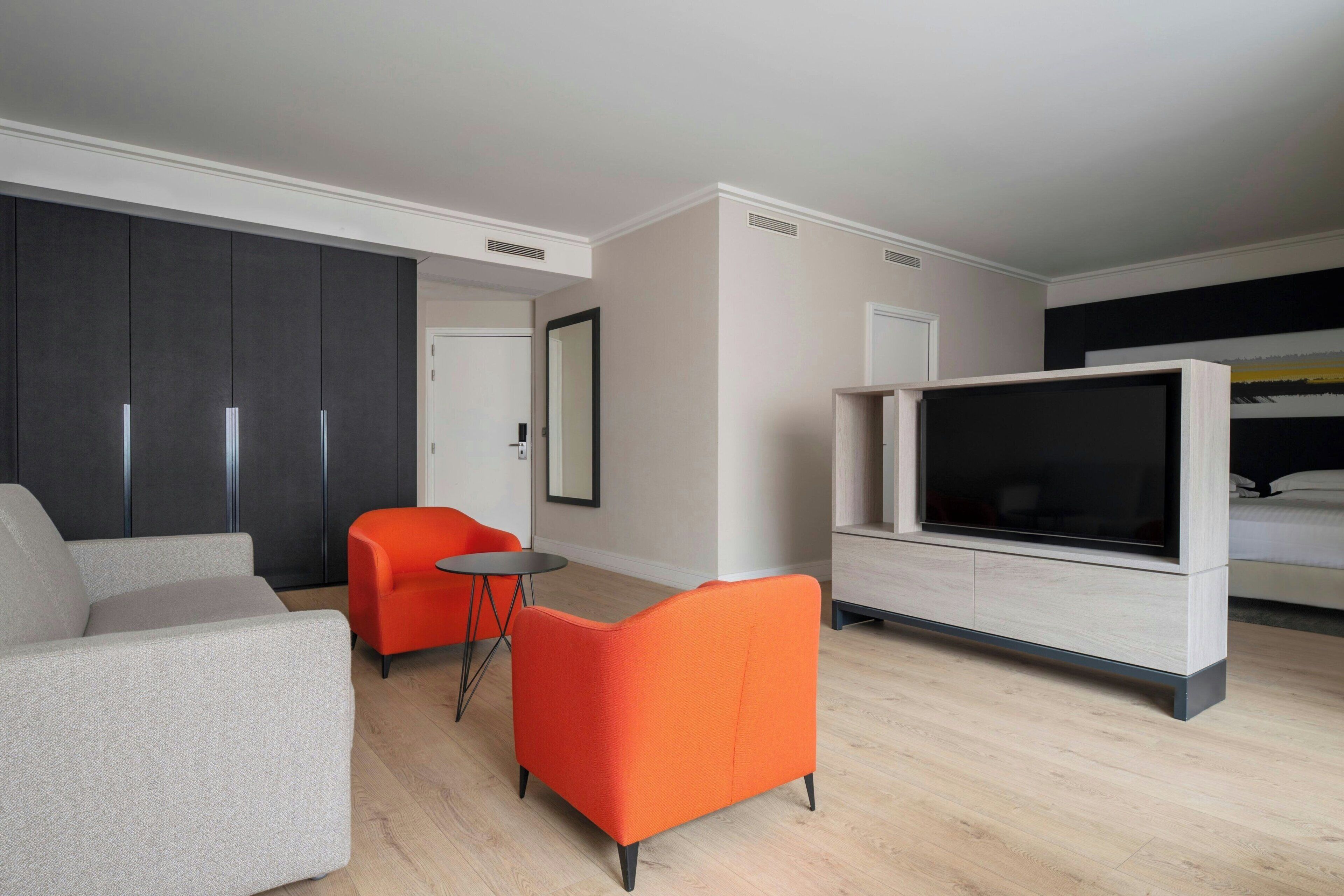 Une chambre d’hôtel moderne avec un canapé, un petit fauteuil orange, une télévision et un lit.