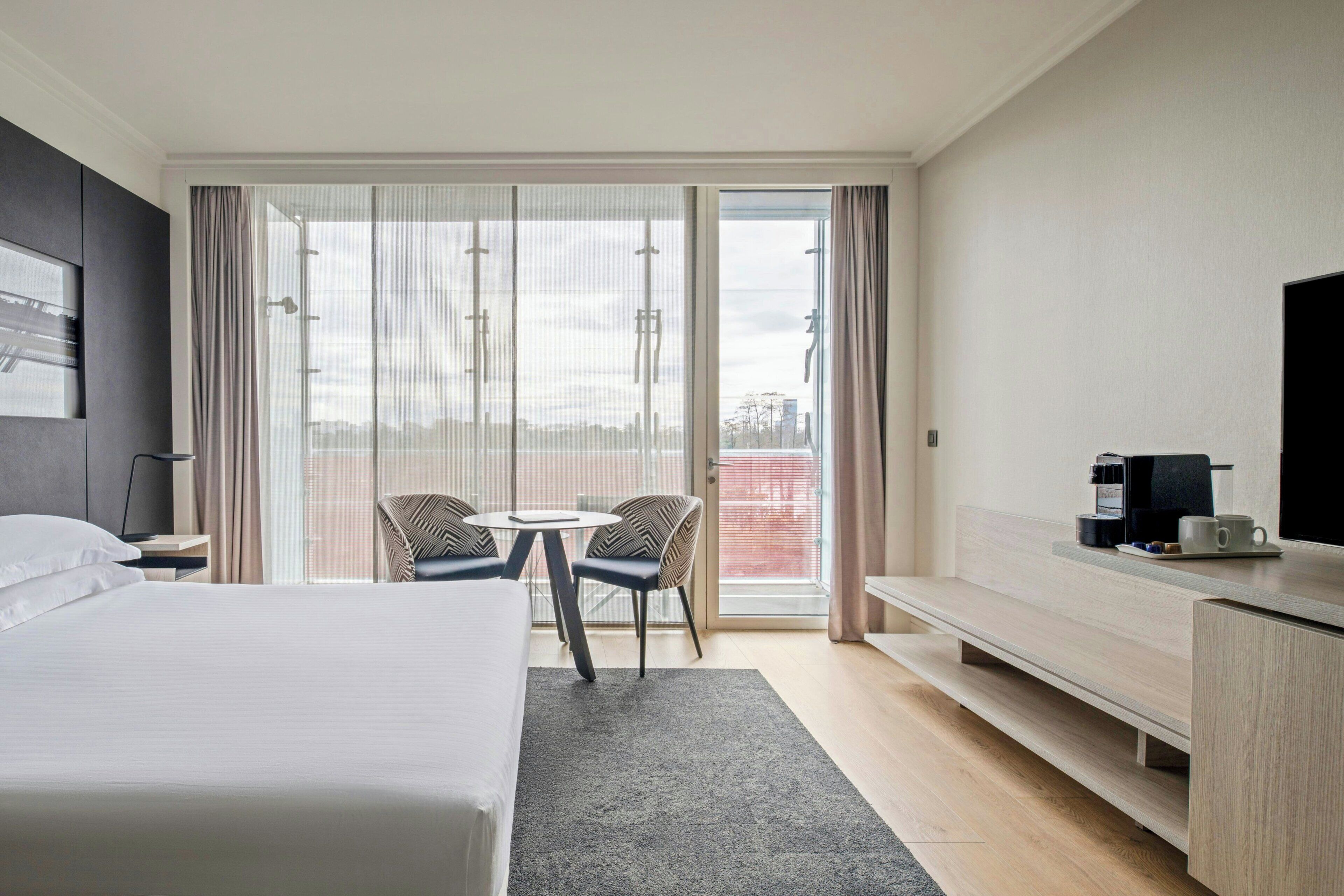 Habitación de hotel con cama, escritorio, silla, mesita y vistas a través de un ventanal grande.