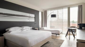 A modern hotel room with a large bed, a smaller bed, a desk with a chair, and a large window with curtains.