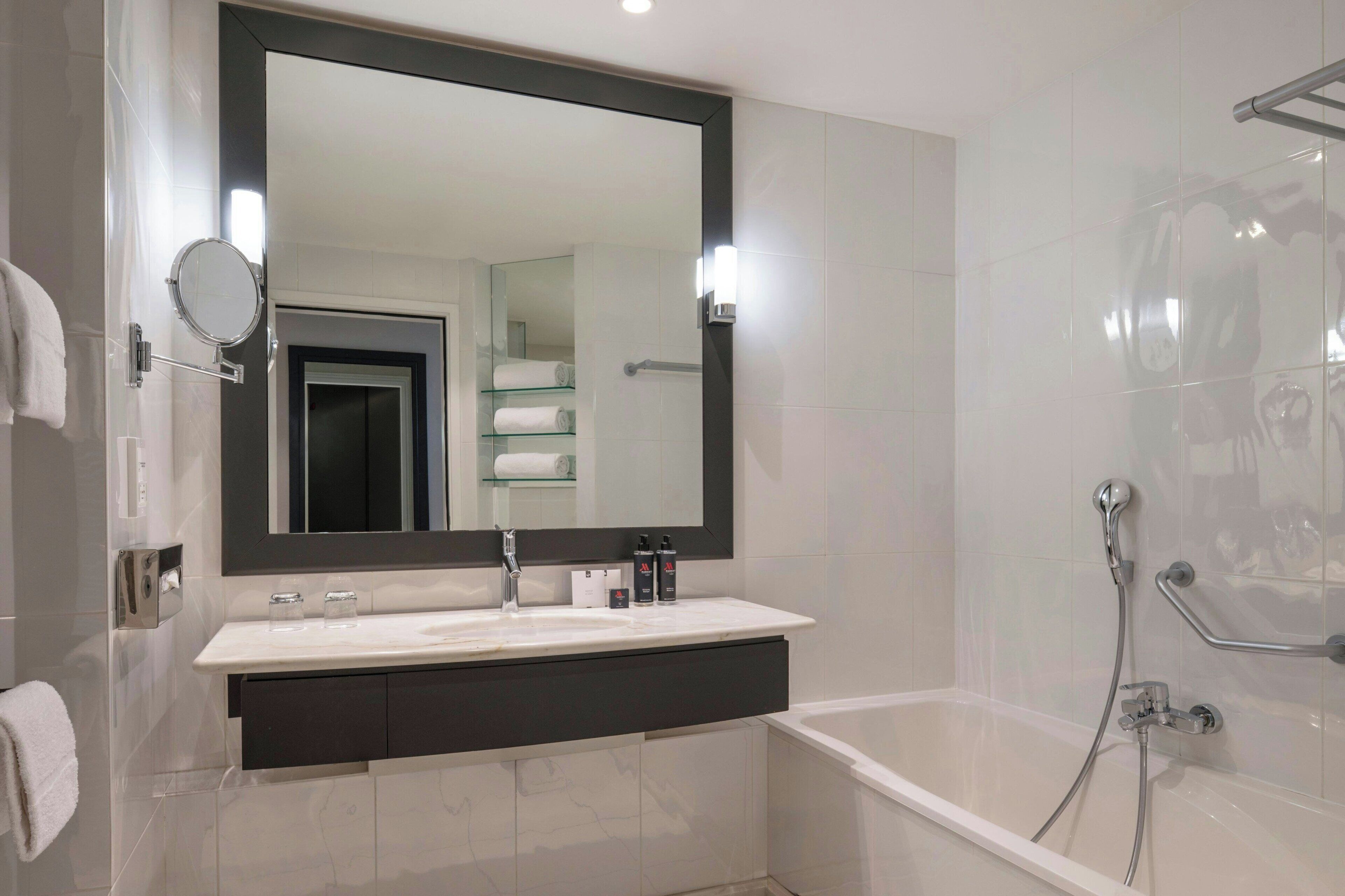 A modern bathroom with a large mirror, white sink, dark vanity, and a bathtub with shower controls.