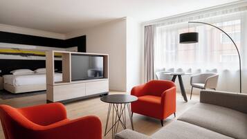 A modern hotel room with a bed, a TV, a dining area, and a large window with curtains.