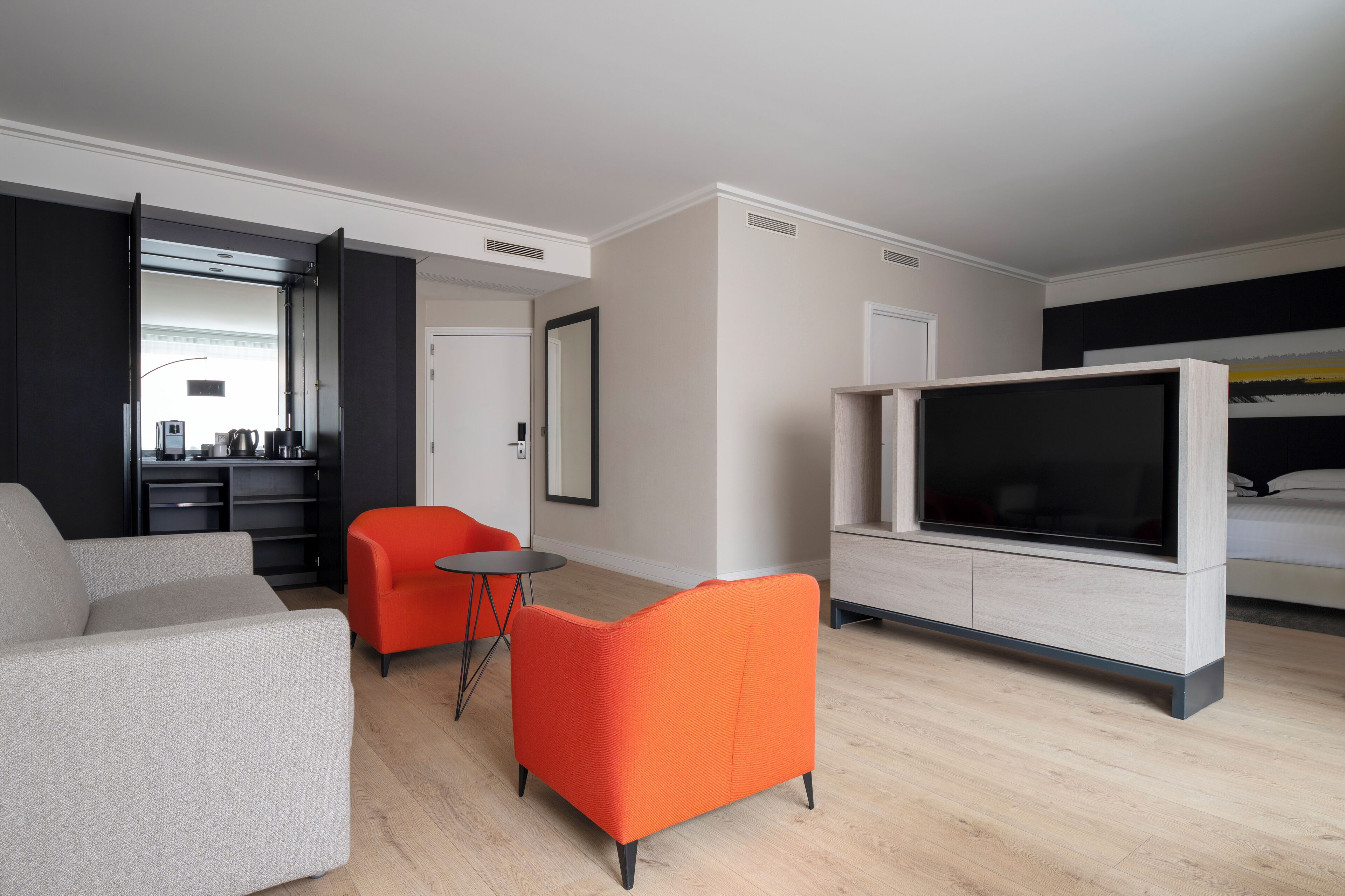 Una habitación de hotel moderna con sofá, un sillón rojo, televisor y un área de comedor con mesa y sillas.
