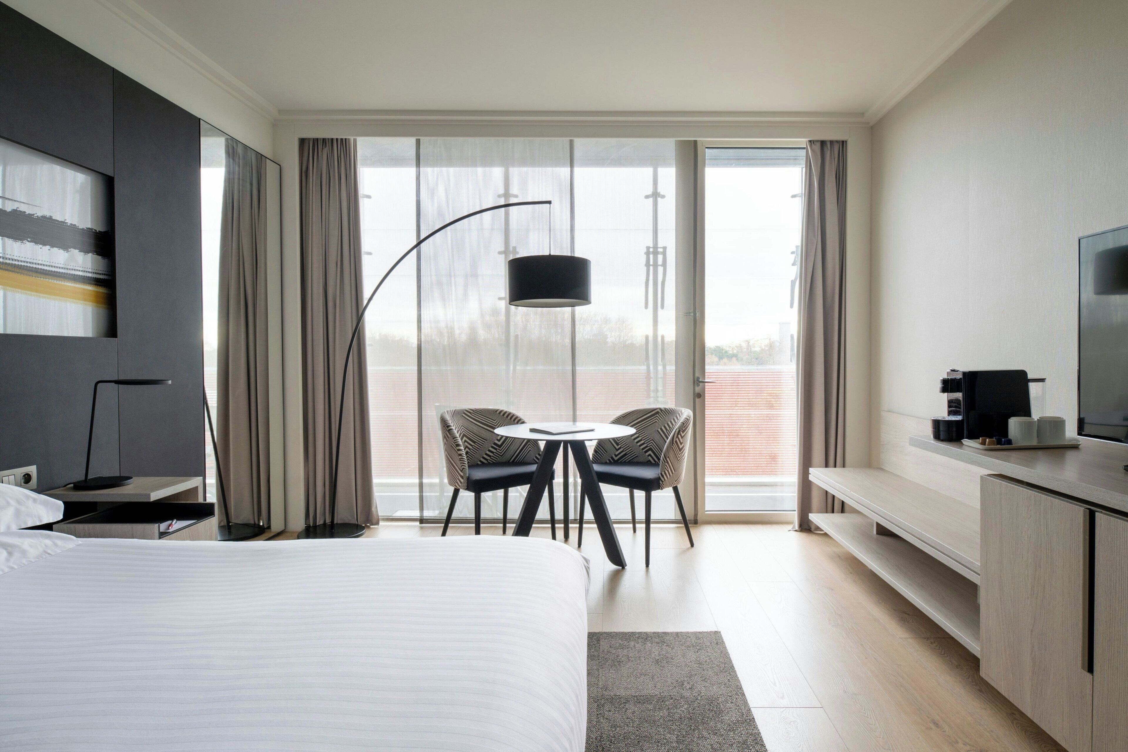 A modern hotel room with a large bed, a desk with a flat-screen TV, a dining area with chairs, and a balcony with curtains.