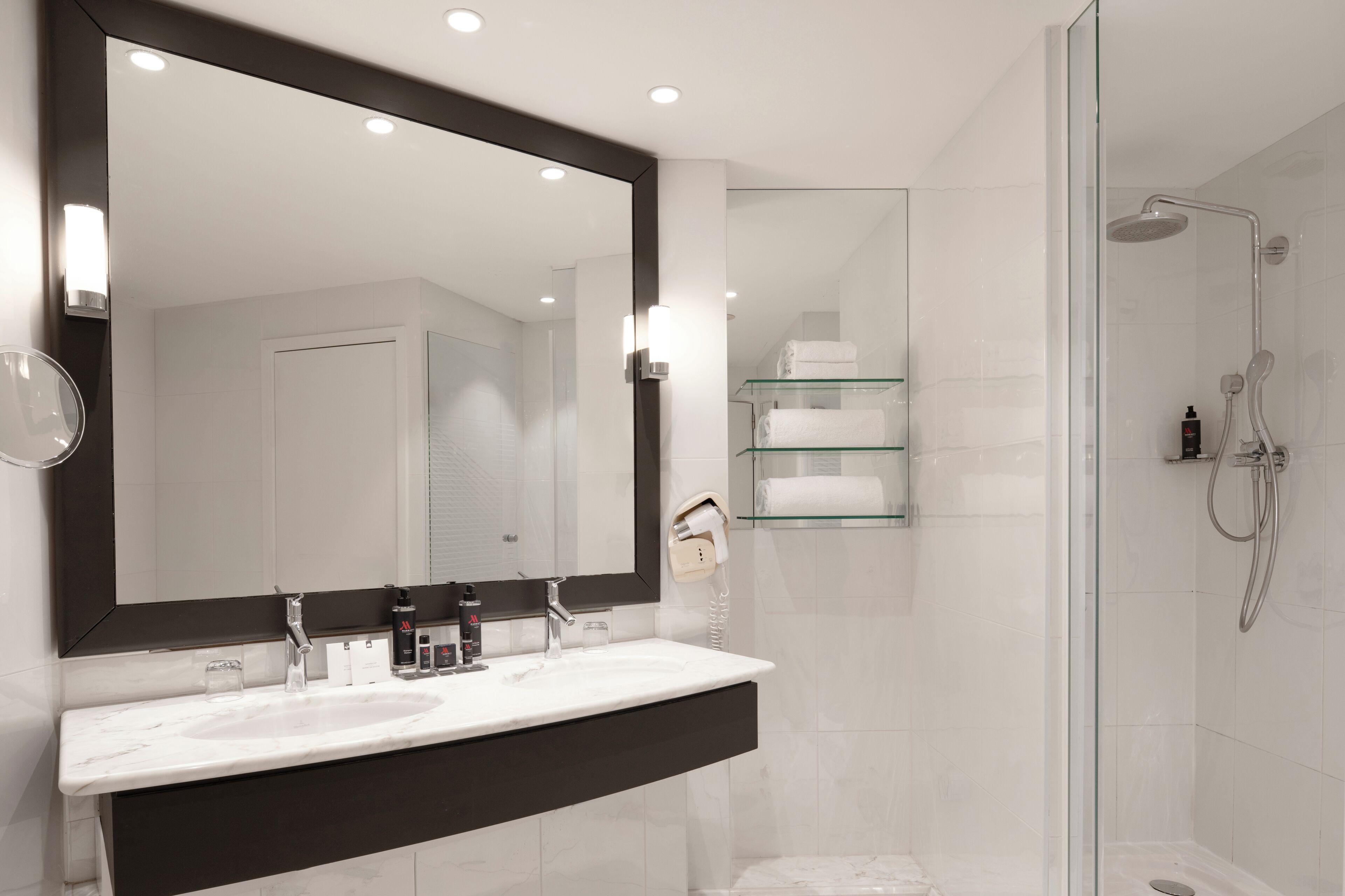 A modern bathroom with a large mirror, two sinks, a glass shower enclosure, and a glass shelf with towels.