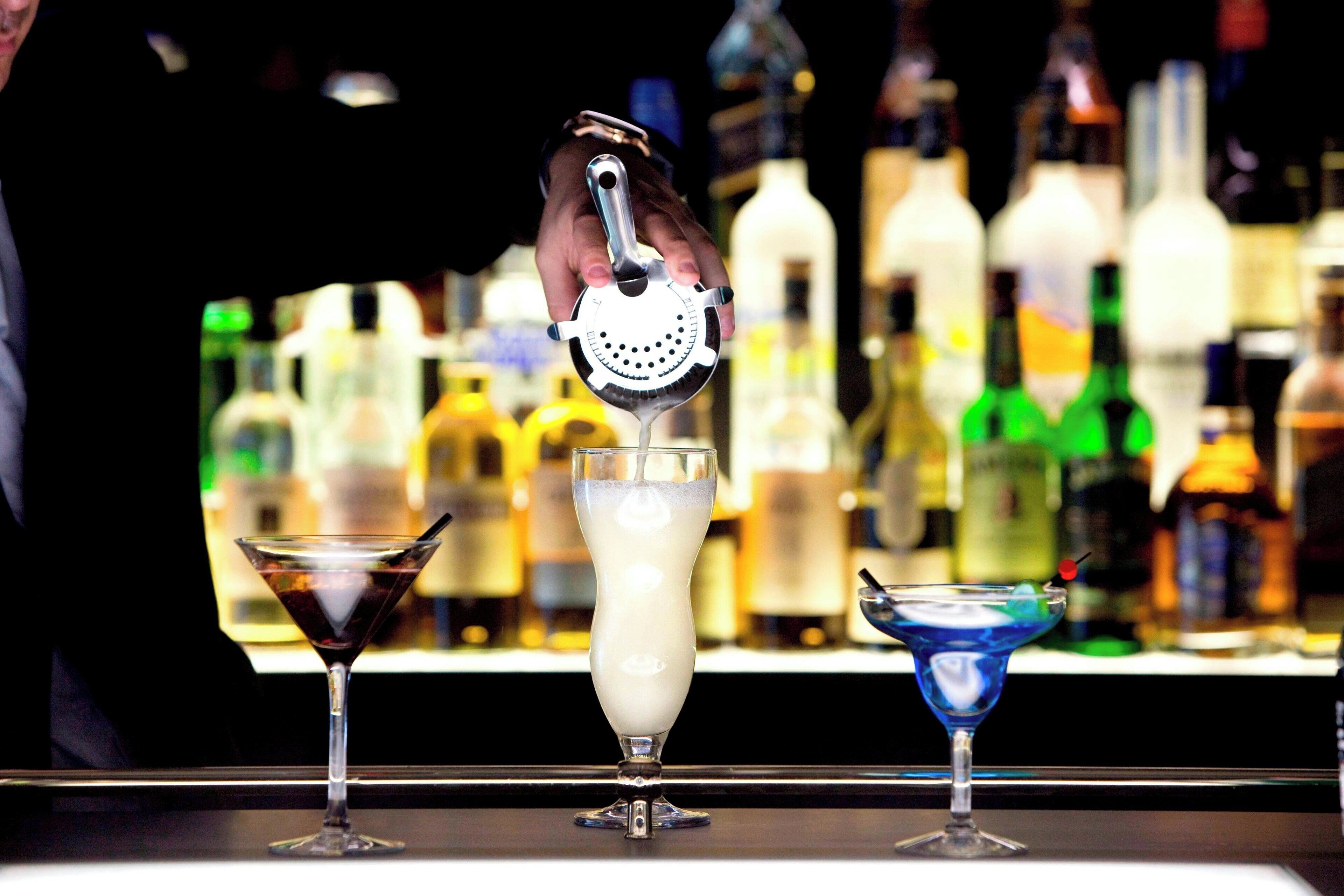 A bar with three cocktails, a bartender's hand, and a variety of bottles in the background.