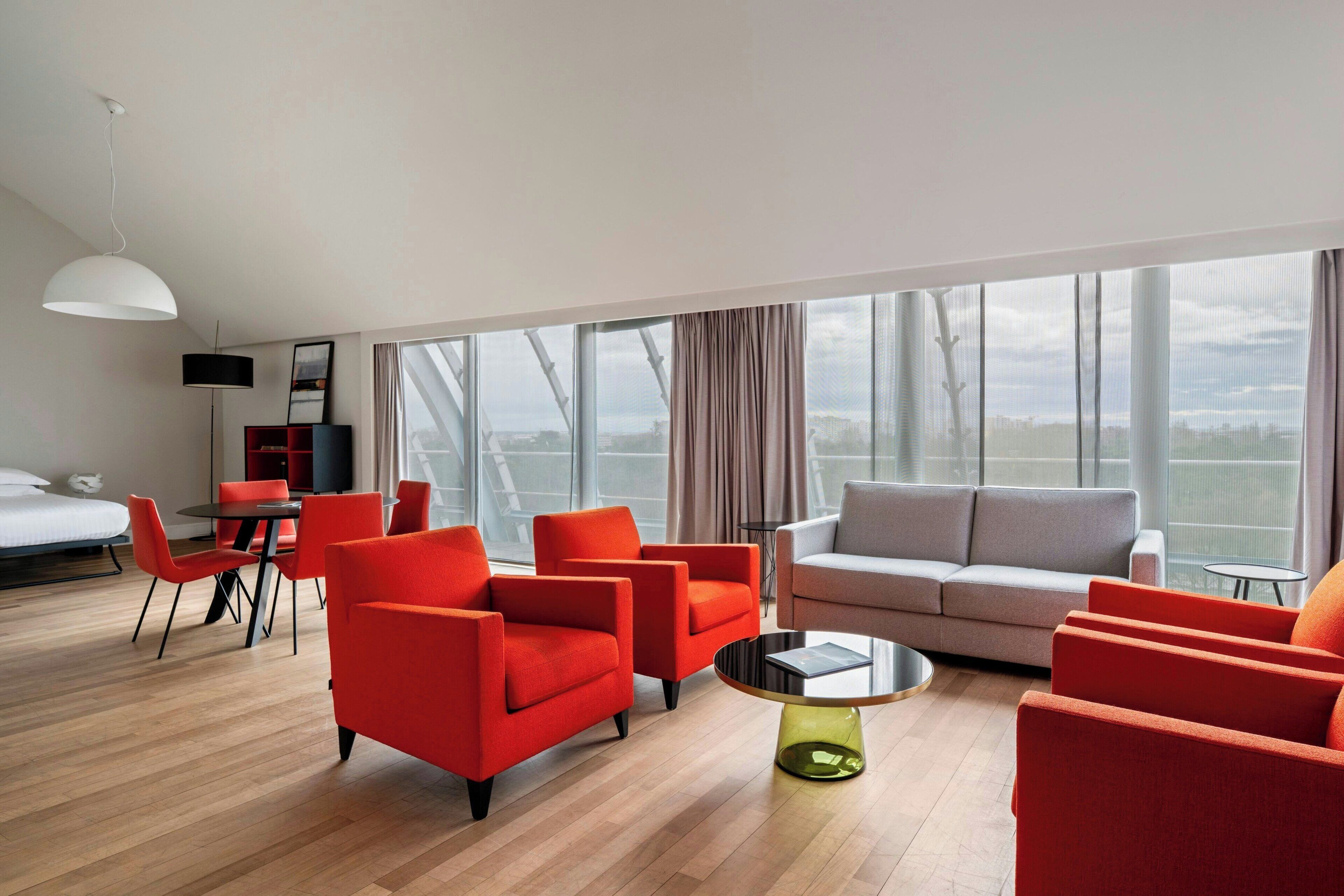 A modern living room with a sofa, red armchairs, a coffee table, and a dining area with chairs and a table.